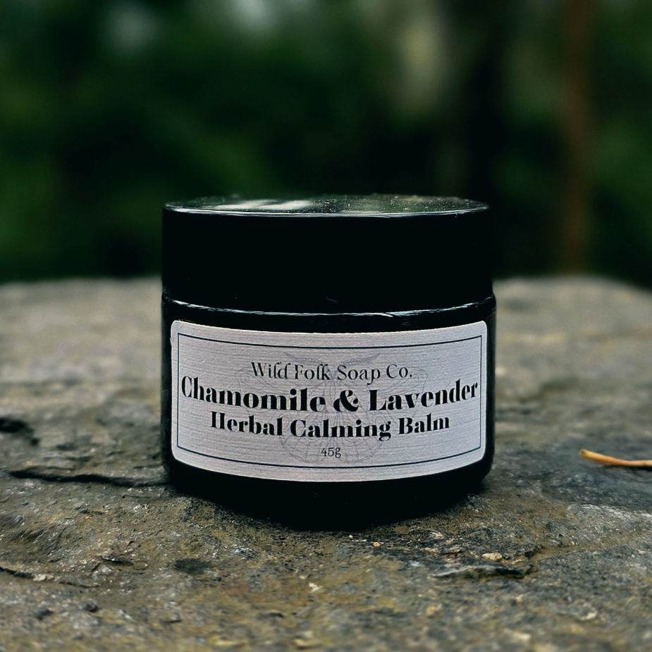 Amber jar of Chamomile & Lavender Herbal Calming Balm by Wild Folk Soap Co. on a stone surface with a blurred green background.
