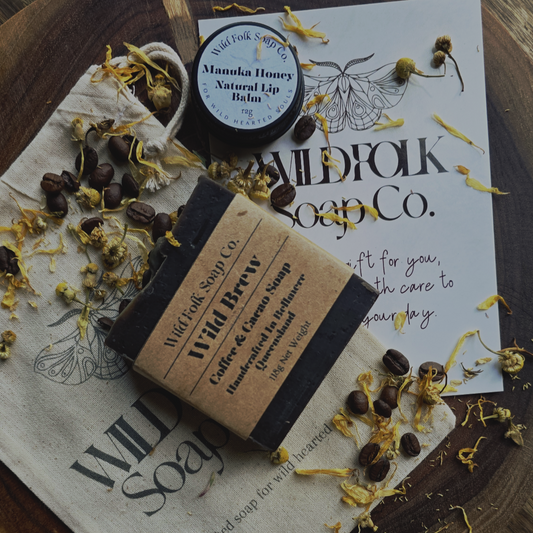 Wild Folk Soap Co. products including a bar of soap and a lip balm on a wooden surface with dried flowers and leaves.