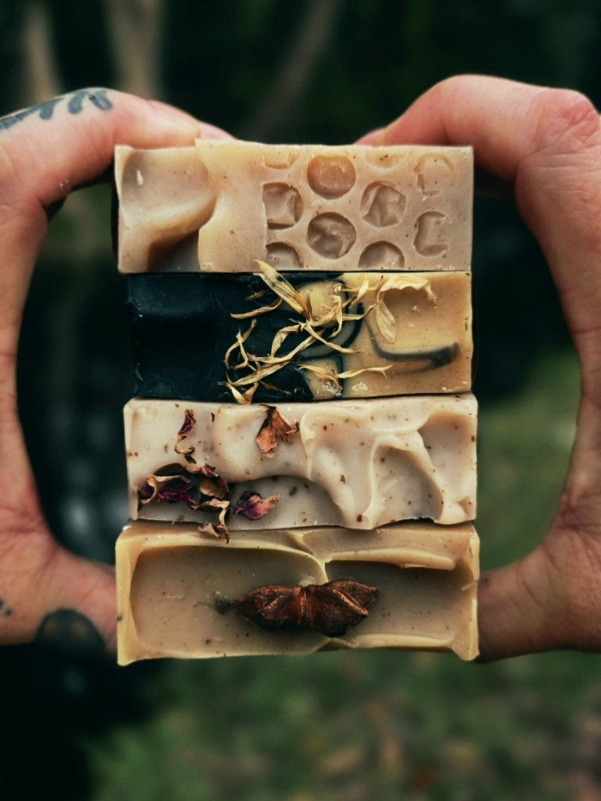 Hand holding five different types of soap bars with natural ingredients against a blurred green background